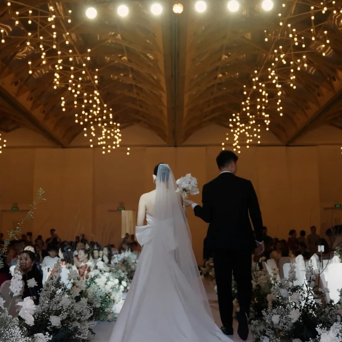 Cô dâu chú rể bước vào ballroom dưới hệ thống đèn trần trang trí.