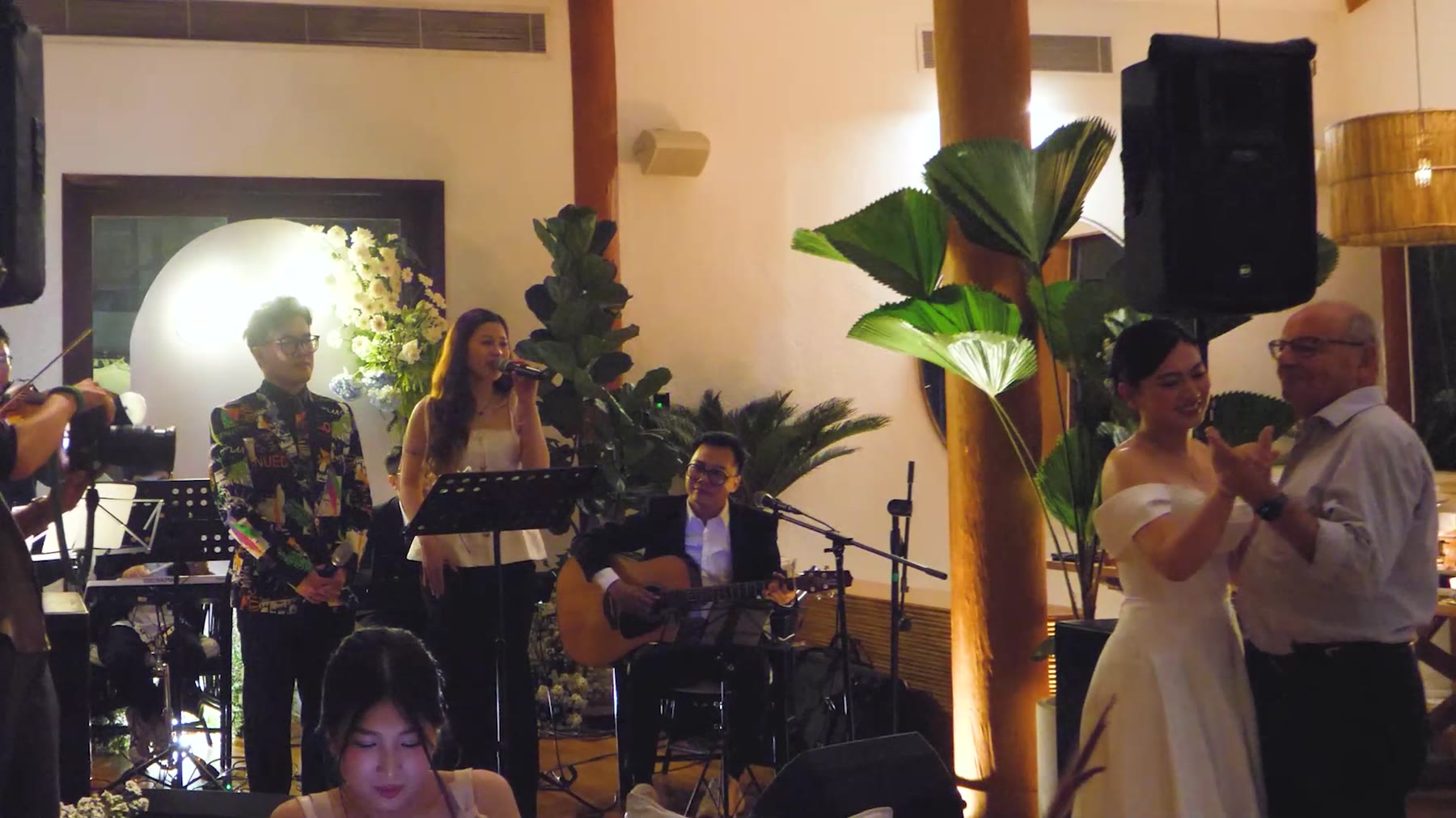 Cô dâu khiêu vũ với bố chú rể trong tiệc cưới khi Acoustic Band Lux Band biểu diễn phía sau.